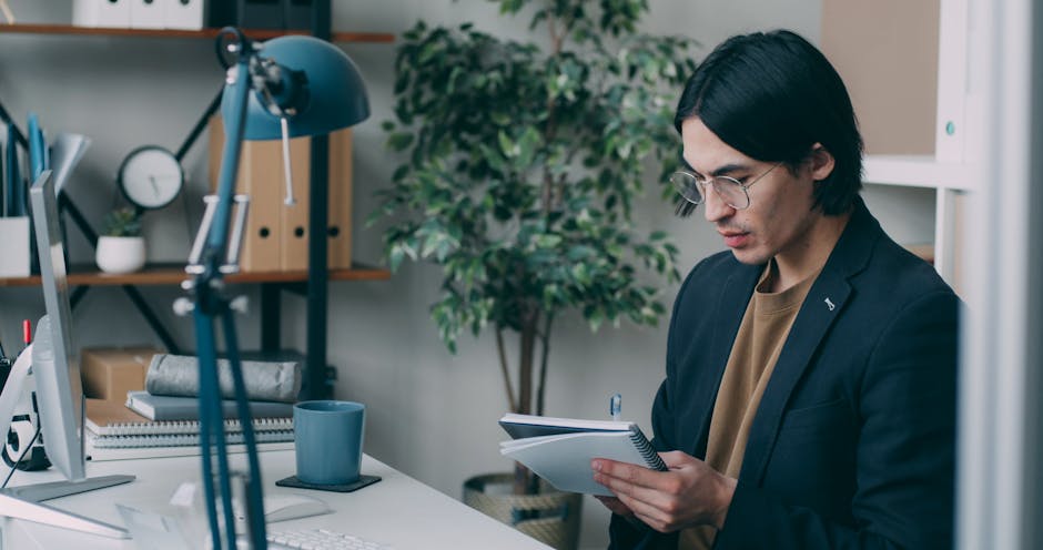 A focused man in a modern office environment, taking notes by his computer desk.