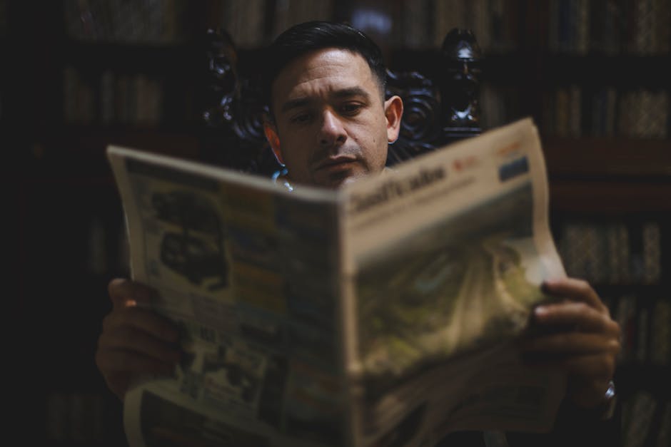 A thoughtful man reads a newspaper in a dimly lit library, conveying solitude and focus.